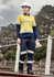 A woman in a yellow and navy work shirt with reflective stripes, wearing a hard hat and gloves, stands outdoors.