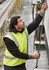 A man in a high-visibility yellow waterproof reversible vest is operating a chain while standing on a gravel surface.