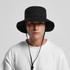 A black wide brim bucket hat with a chin strap, showcased on a model wearing a white sweatshirt.