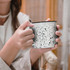 A ceramic mug featuring a speckled black and white design, held by a person with one hand. The mug has a contrasting rim.