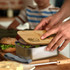 A stainless steel lunch box with a bamboo lid, filled with sandwiches and greens, on a wooden surface.