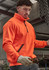 An orange unisex 1/4 zip pullover is worn by a person inside a workshop, featuring protective glasses and gloves.
