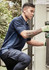 A man wearing a navy blue short sleeve polo with a patterned side panel is kneeling near equipment outdoors.
