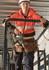 A unisex hi-vis full zip stretch taped hoodie in bright orange, worn by a person holding a power tool while on scaffolding.