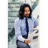 A man with long hair wearing a blue and white striped button-up shirt and dark tie, reading a newspaper.