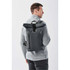 A grey roll-top backpack is worn by a man, showing a zippered front pocket and featuring a logo.