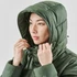 A woman wearing a green puffer jacket with a hood, adjusting it around her face. The jacket has a subtle logo.