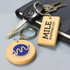 A wooden rectangle key ring with a logo, accompanied by a round key ring, resting on a surface with keys and a notebook.