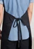 A bib apron in black, featuring a tie at the back and worn over a light blue shirt.