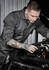A man wearing a dark grey overall inspects the engine of a car, showcasing detailed tattoos on his arm.