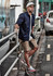 A man in a navy shirt and khaki shorts stands by a railing, with a skateboard and cars in the background.