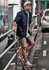 A man wearing a navy shirt and beige stretch shorts stands by a railing, with a skateboard beside him.