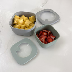 A set of two square food storage containers, one light gray and one pistachio green, are shown on a marble surface with their see-through lids off, filled with fresh pineapple and strawberries. A set of two square food storage containers, one light gray and one pistachio green, are shown on a marble surface with their see-through lids off, filled with fresh pineapple and strawberries.