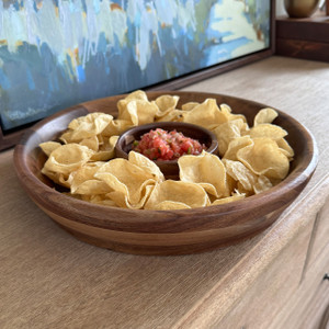 A large, round acacia wood chip and dip serving platter is displayed on a wooden table, filled with tortilla chips that surround the integrated center bowl of fresh salsa.