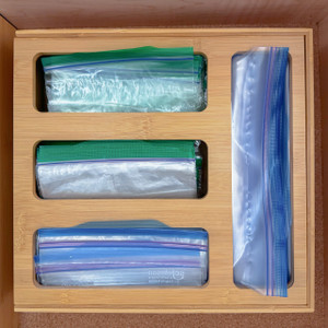 A top-down view of a bamboo bag organizer placed inside a kitchen drawer, with its four compartments neatly filled with gallon, quart, sandwich, and snack-sized plastic storage bags. A top-down view of a bamboo bag organizer placed inside a kitchen drawer, with its four compartments neatly filled with gallon, quart, sandwich, and snack-sized plastic storage bags.