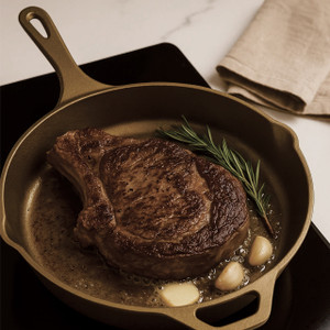 Golden cast iron skillet searing a ribeye with garlic and rosemary on a stovetop; helper handle and pour spout visible. Golden cast iron skillet searing a ribeye with garlic and rosemary on a stovetop; helper handle and pour spout visible.
