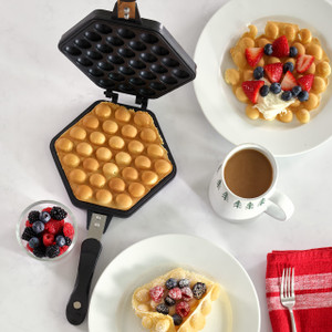 A complete breakfast scene featuring an open Hong Kong egg waffle pan with a freshly cooked golden bubble waffle inside, surrounded by plated waffles topped with fresh berries and cream, and a cup of coffee.