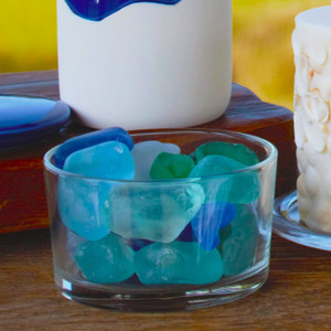 A clear glass container filled with beautiful blue, green, and frosted sea glass scented stones is displayed as a decorative piece on a wooden table.
