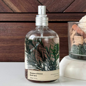 A decorative Bergamot & Botanicals room spray in a clear glass bottle, featuring beautiful botanical and cinnamon stick inclusions, is elegantly styled against a dark wood background.