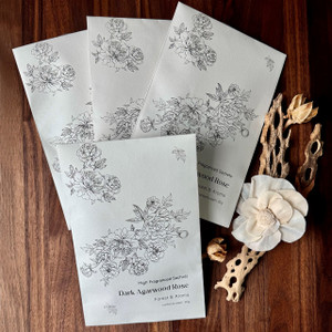 A set of four elegant Dark Agarwood Rose scented sachets, featuring a beautiful black floral line drawing, are artfully arranged on a dark wood table with decorative dried botanicals.