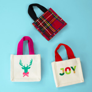 Trio of mini gift totes on blue background—red tartan plaid, white “JOY,” and white deer head design Trio of mini gift totes on blue background—red tartan plaid, white “JOY,” and white deer head design