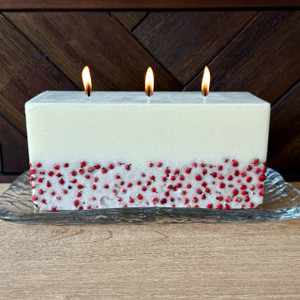 Rectangular white soy wax candle with embedded red berries and three lit wicks, displayed on a clear glass tray. Rectangular white soy wax candle with embedded red berries and three lit wicks, displayed on a clear glass tray.