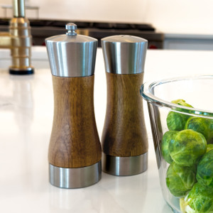 Stainless and acacia salt shaker and pepper grinder set standing on a kitchen counter beside a bowl of Brussels sprouts.