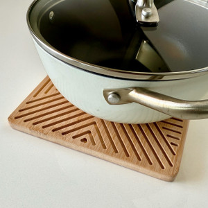 Saucepan resting on a square bamboo laser-cut trivet with geometric grooves on a countertop. Saucepan resting on a square bamboo laser-cut trivet with geometric grooves on a countertop.