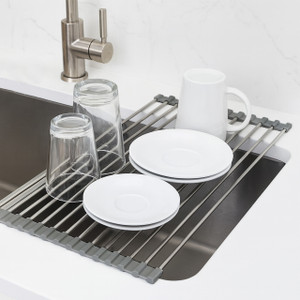 Stainless roll-up dish drying rack across the sink holding plates and glasses, gray silicone ends for secure grip.