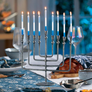 The elegant, dark iron Liora Menorah is shown as a centerpiece on a beautifully set Hanukkah dinner table, with all nine blue and white taper candles lit and glowing. The elegant, dark iron Liora Menorah is shown as a centerpiece on a beautifully set Hanukkah dinner table, with all nine blue and white taper candles lit and glowing.