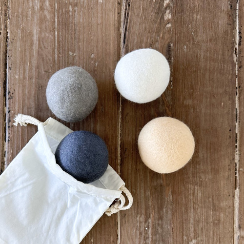 A set of four wool dryer balls in assorted neutral colors (white, cream, light gray, and dark gray) are displayed on a rustic wood surface, spilling out of their white cotton drawstring storage pouch.