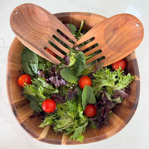 A pair of beautifully crafted acacia wood salad servers, shaped like rounded hands, are tossed in a large wooden bowl filled with fresh mixed greens and cherry tomatoes.