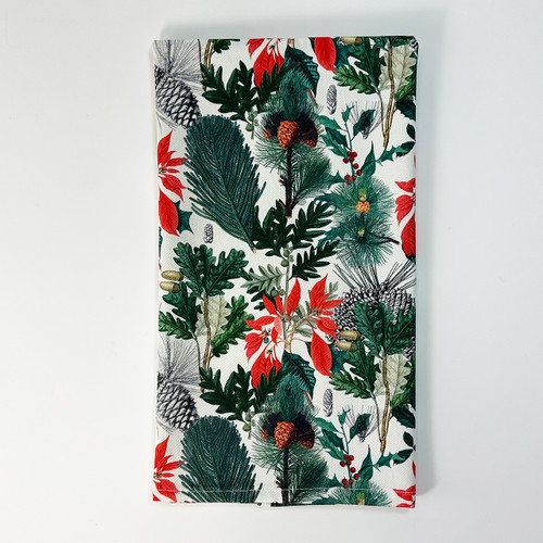 A neatly folded white cotton hand towel featuring a festive "Botanical Jubilee" all-over print of green pine branches, pinecones, and red poinsettia flowers, standing upright against a clean white background.