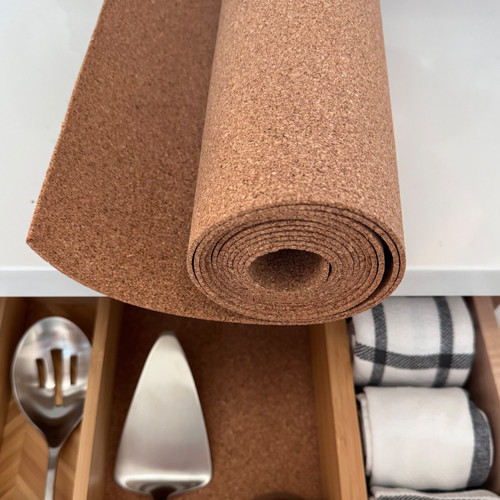 A roll of natural cork drawer liner is partially unrolled on a white countertop, positioned above an open kitchen drawer that has been lined with the same cork material, neatly organizing serving utensils and dish towels.