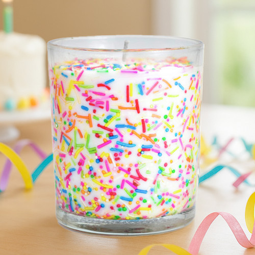 : A close-up lifestyle shot of the Celebration Sprinkles candle, showing the vibrant confetti-like sprinkles in the wax, with a birthday cake and lit candle visible in the blurred background.