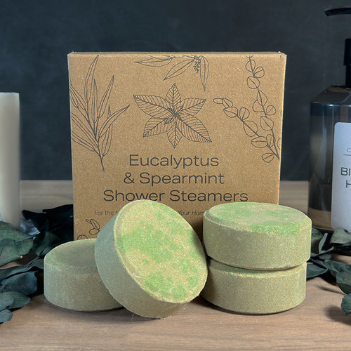 Four round, green Eucalyptus & Spearmint shower steamers are elegantly displayed in a spa-like setting, with some tablets stacked and others resting in front of their natural kraft paper gift box.