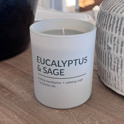 A Eucalyptus & Sage scented soy candle in a minimalist, frosted white glass jar is styled on a wooden table, creating a serene and relaxing home atmosphere.
