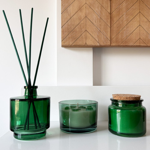 An elegant Green Glass Diffuser & Candles Set, featuring a reed diffuser, an apothecary candle with a cork lid, and a stones inclusion candle, is beautifully arranged on a modern white shelf.