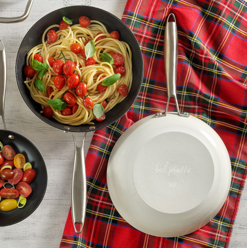 Overhead of two Bel Piatto No.10 cast iron fry pans—one filled with cherry-tomato pasta, the other flipped to show the glossy white enameled exterior and stainless handle on a plaid towel