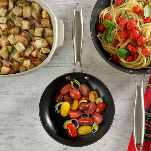 An 8-inch cast iron fry pan with a nonstick ceramic interior, containing sautéed cherry tomatoes and leeks, next to a casserole dish filled with roasted bread and vegetables. The pan has a stainless steel handle and is placed on a white surface with a plaid cloth.