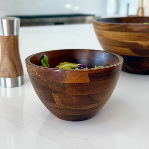An acacia wood individual bowl filled with a fresh, colorful salad on a white kitchen countertop. The bowl displays its distinctive wood grain and tapered shape, with a blurred wooden pepper mill and another bowl in the background.