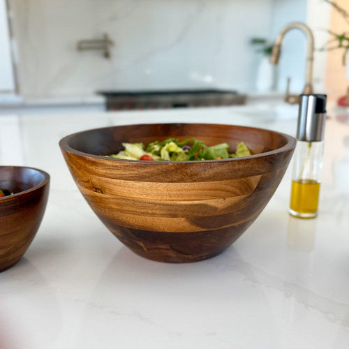 Acacia large serving bowl holding a fresh green salad on a kitchen counter beside a glass oil bottle.