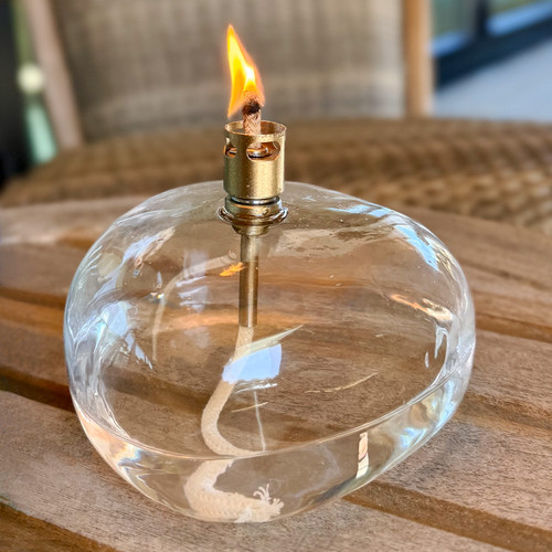 An elegant, clear glass table top torch with a lit flame is sitting on an outdoor wooden patio table, creating a warm and inviting ambiance.