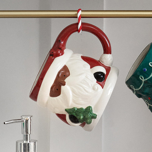 A whimsical, hand-painted ceramic mug shaped like Santa Claus's head, with a small Christmas tree and a red cardinal, is shown hanging by its large, festive handle from a gold bar.