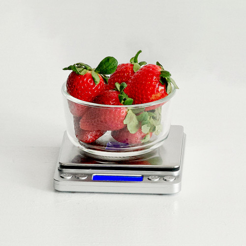Glass bowl of fresh strawberries on a compact stainless steel digital kitchen scale with blue backlit display on a white surface