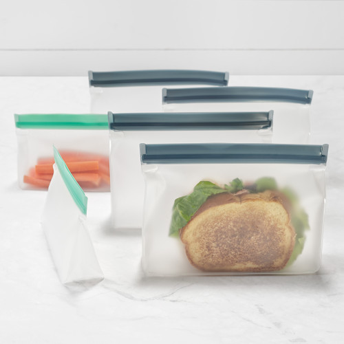 A set of reusable, translucent zip pouches are displayed on a white marble surface. One pouch in the foreground holds a sandwich, another contains carrot sticks, demonstrating their use as an eco-friendly alternative to disposable plastic bags.