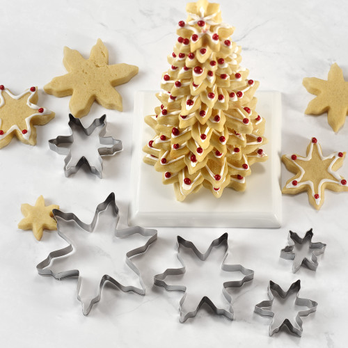 A festive holiday baking scene featuring a set of five stainless steel snowflake cookie cutters in graduated sizes, displayed next to baked sugar cookies and an impressive, edible Christmas tree made from stacked snowflake cookies.