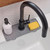 A gray silicone faucet mat is fitted neatly around the base of a modern black faucet on a white kitchen sink, holding a soap dispenser and a yellow sponge, keeping the area clean and dry.