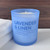 A Lavender & Linen scented soy candle in a tranquil, frosted light blue glass jar is displayed next to a soft, folded white towel, evoking a sense of calm and relaxation.
