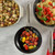 An 8-inch cast iron fry pan with a nonstick ceramic interior, containing sautéed cherry tomatoes and leeks, next to a casserole dish filled with roasted bread and vegetables. The pan has a stainless steel handle and is placed on a white surface with a plaid cloth.